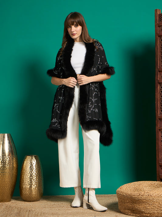 Model wearing black faux fur-trimmed shawl with crystal vine motifs draped over white top and trousers