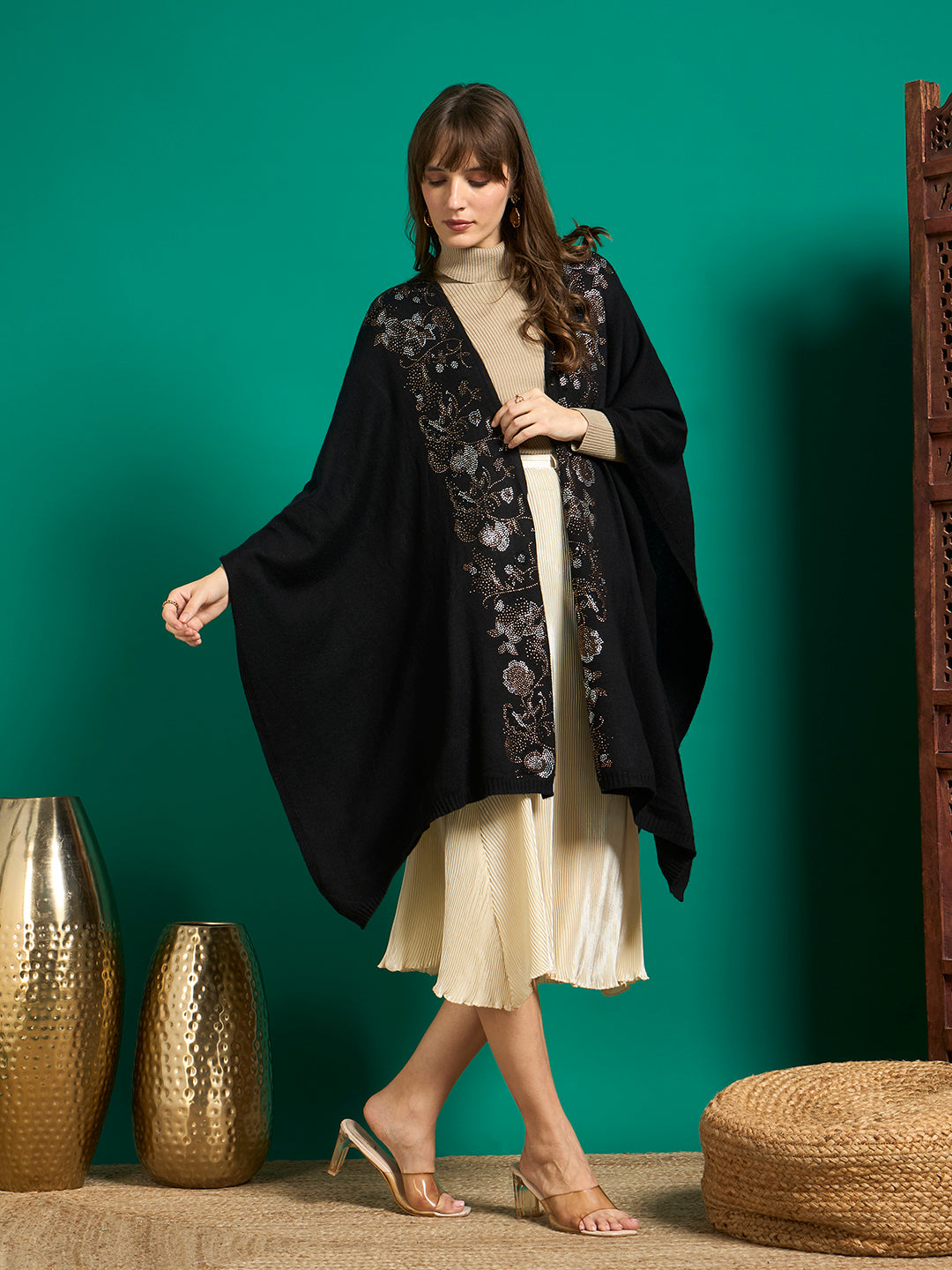 Black embellished cape with draped batwing sides worn over a beige turtleneck and cream pleated skirt