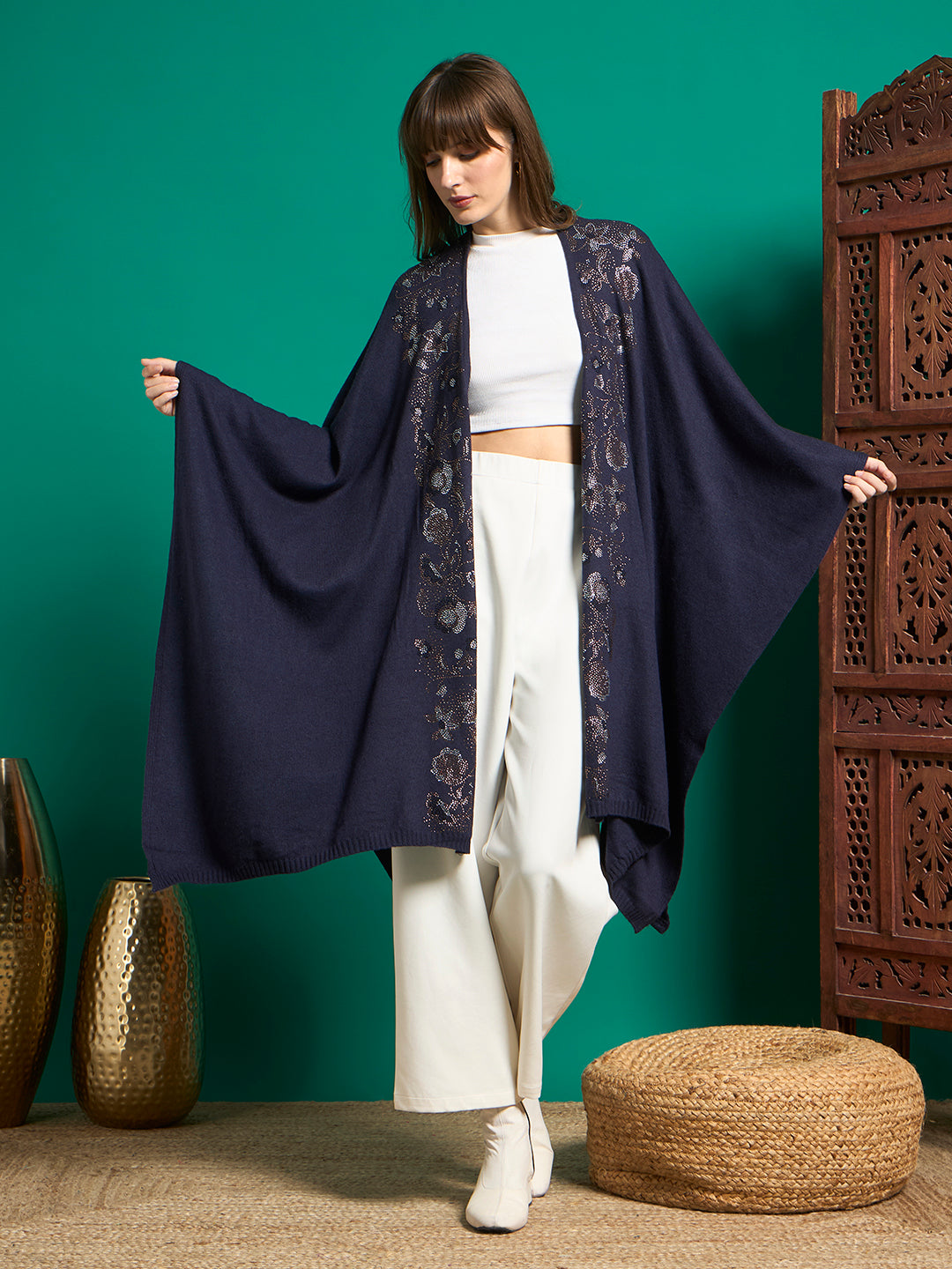 Model in navy knit cape with floral stud embellishment on front panels, arms spread to show batwing drape